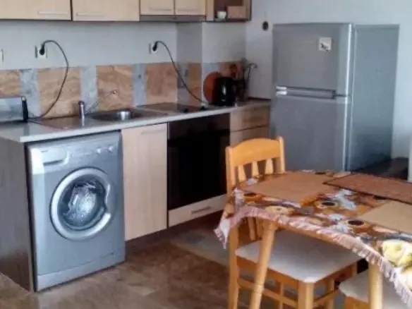 Современная кухня с деревянными шкафчиками, стиральной машиной, холодильником и обеденным столом в квартире в Болгарии.