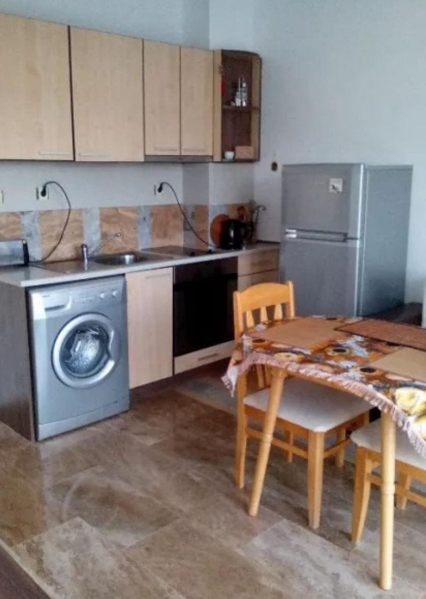 Современная кухня с деревянными шкафчиками, стиральной машиной, холодильником и обеденным столом в квартире в Болгарии.