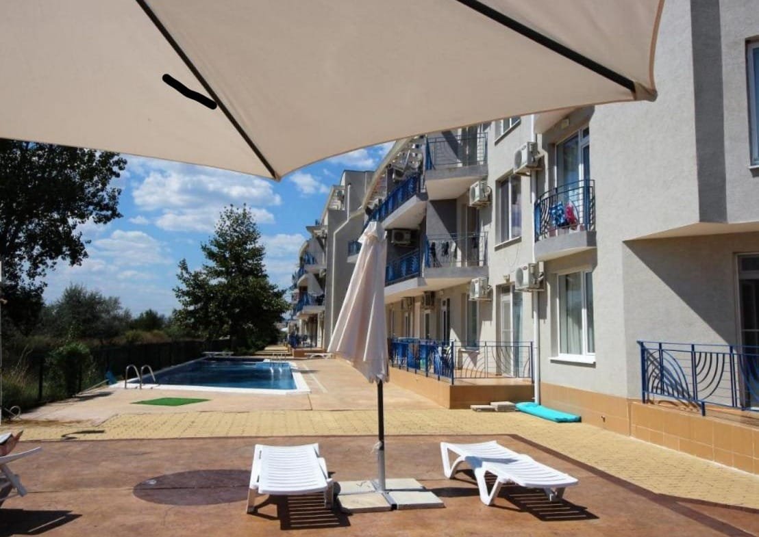 Светлый комплекс недвижимости в Болгарии с бассейном, зонтиками и шезлонгами, окруженный зелеными насаждениями.