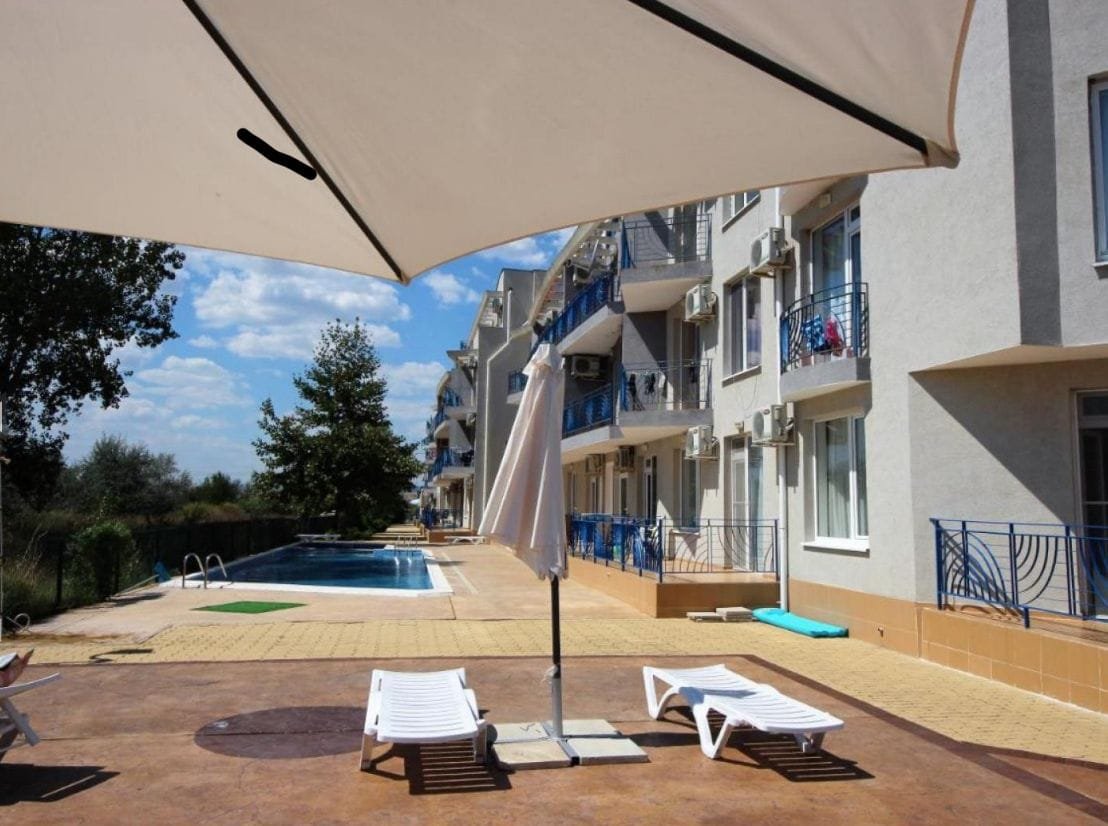 Светлый комплекс недвижимости в Болгарии с бассейном, зонтиками и шезлонгами, окруженный зелеными насаждениями.