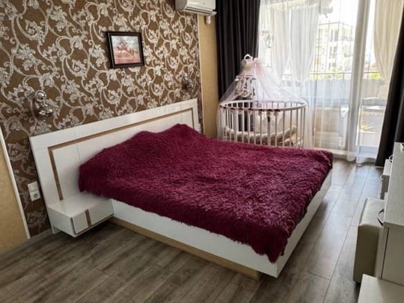 Современная спальня с кроватью и уютным покрывалом, окнами с видом на улицу и детской кроваткой в углу.