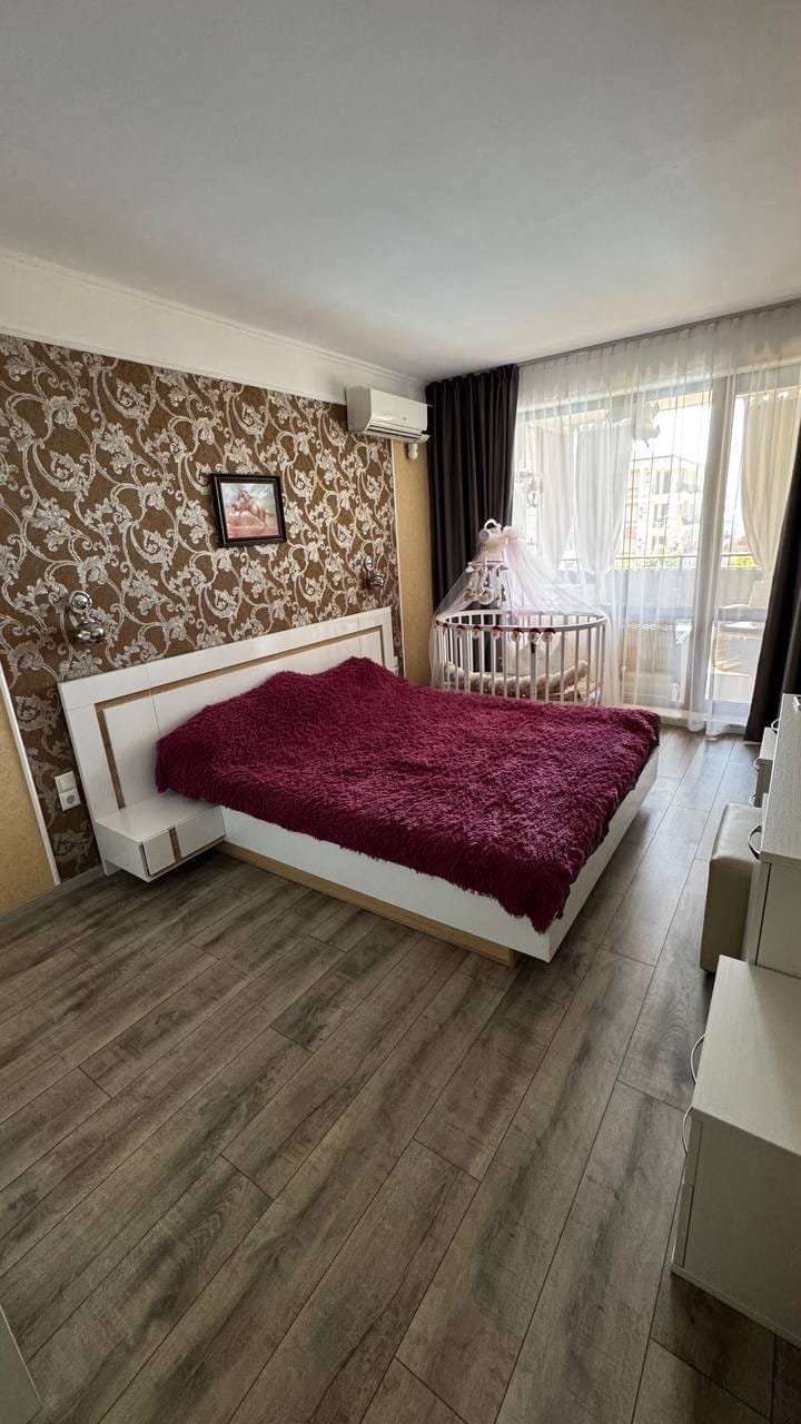 Современная спальня с кроватью и уютным покрывалом, окнами с видом на улицу и детской кроваткой в углу.