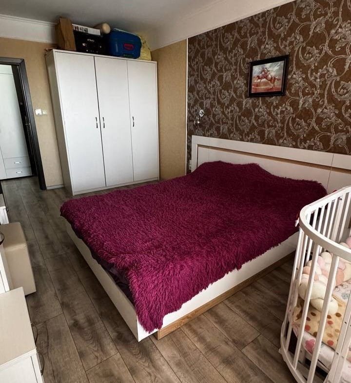 Современная спальня с двуспальной кроватью, светлыми стенами, деревянным полом и детской кроваткой. Идеально для покупки недвижимости в Болгарии.