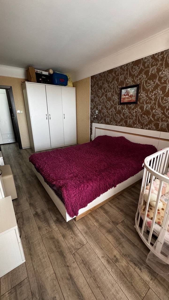 Современная спальня с двуспальной кроватью, светлыми стенами, деревянным полом и детской кроваткой. Идеально для покупки недвижимости в Болгарии.