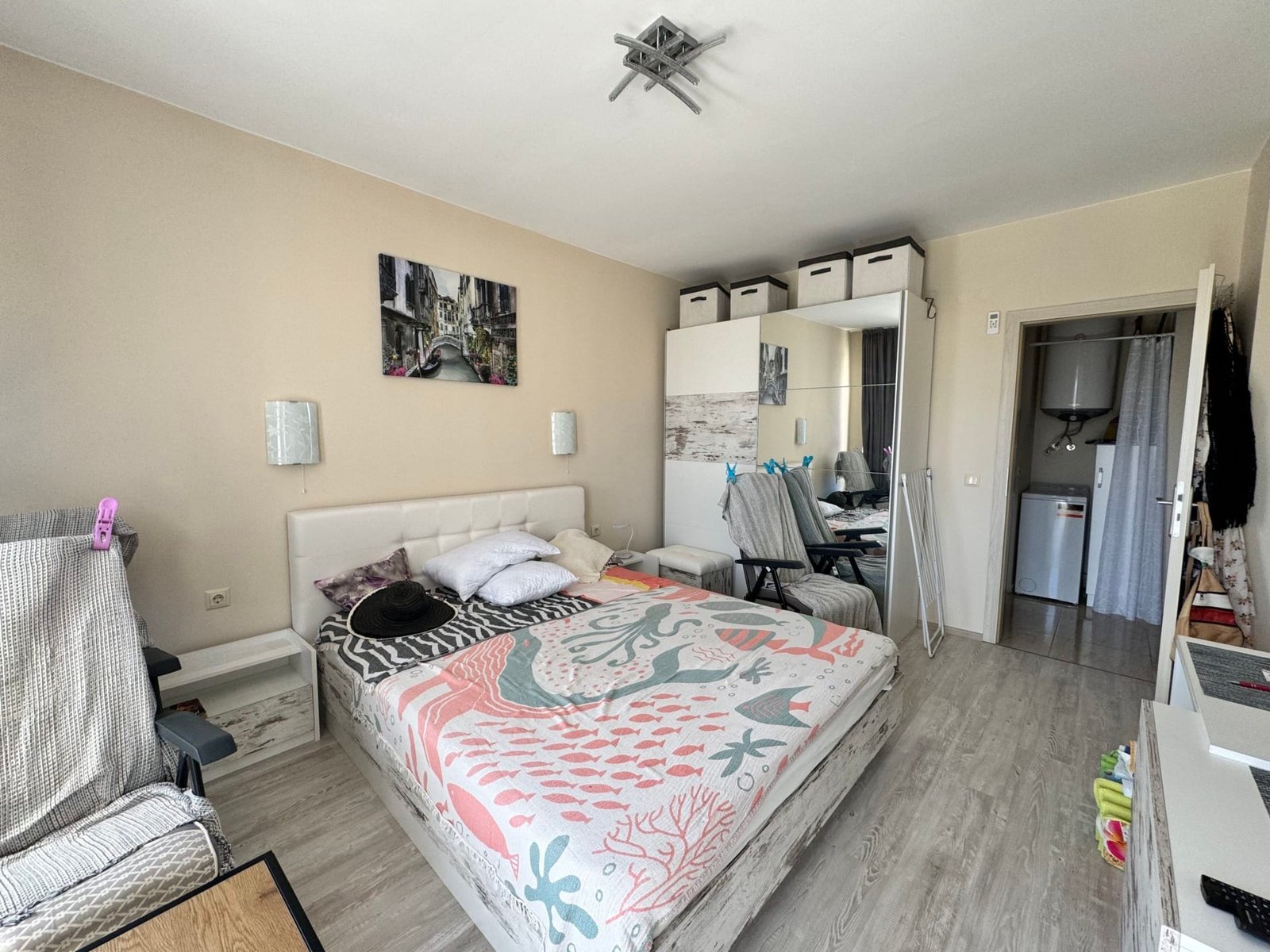 Современная спальня с кроватью и стильным декором Уютная спальня с двуспальной кроватью, светлыми стенами и современным интерьером, идеальна для покупки недвижимости в Болгарии.