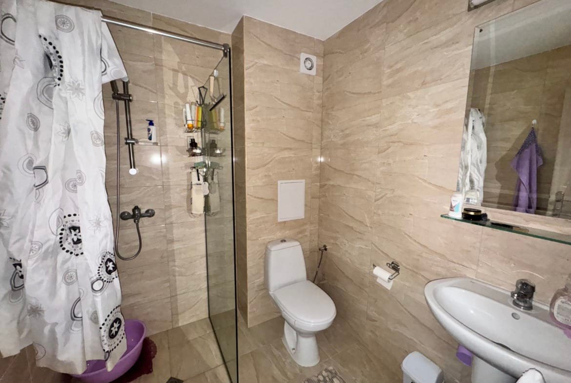 Светлая ванная комната с душевой кабиной, унитазом и раковиной. Стены отделаны светлой плиткой, в углу — гладильная доска.