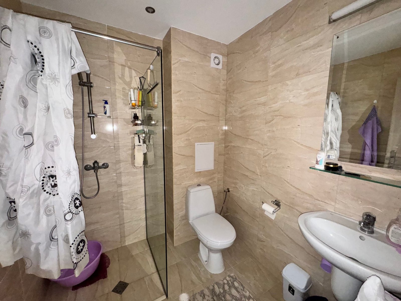 Современная ванная комната с душем и сантехникой Светлая ванная комната с душевой кабиной, унитазом и раковиной. Стены отделаны светлой плиткой, в углу — гладильная доска.