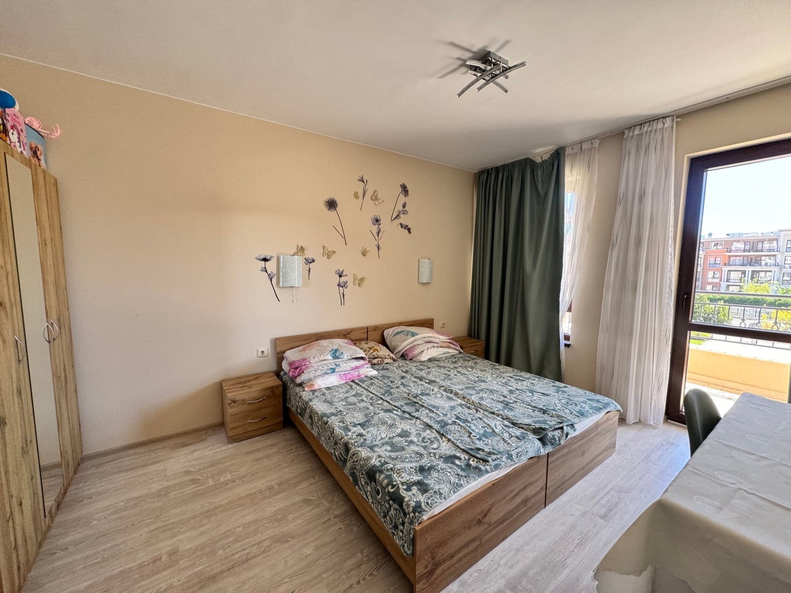 Уютная спальня с двумя кроватями и большим окном. Современная спальня с двуспальной кроватью, шкафом и большим окном, оформленная в нежных тонах, идеальна для отдыха в Болгарии.