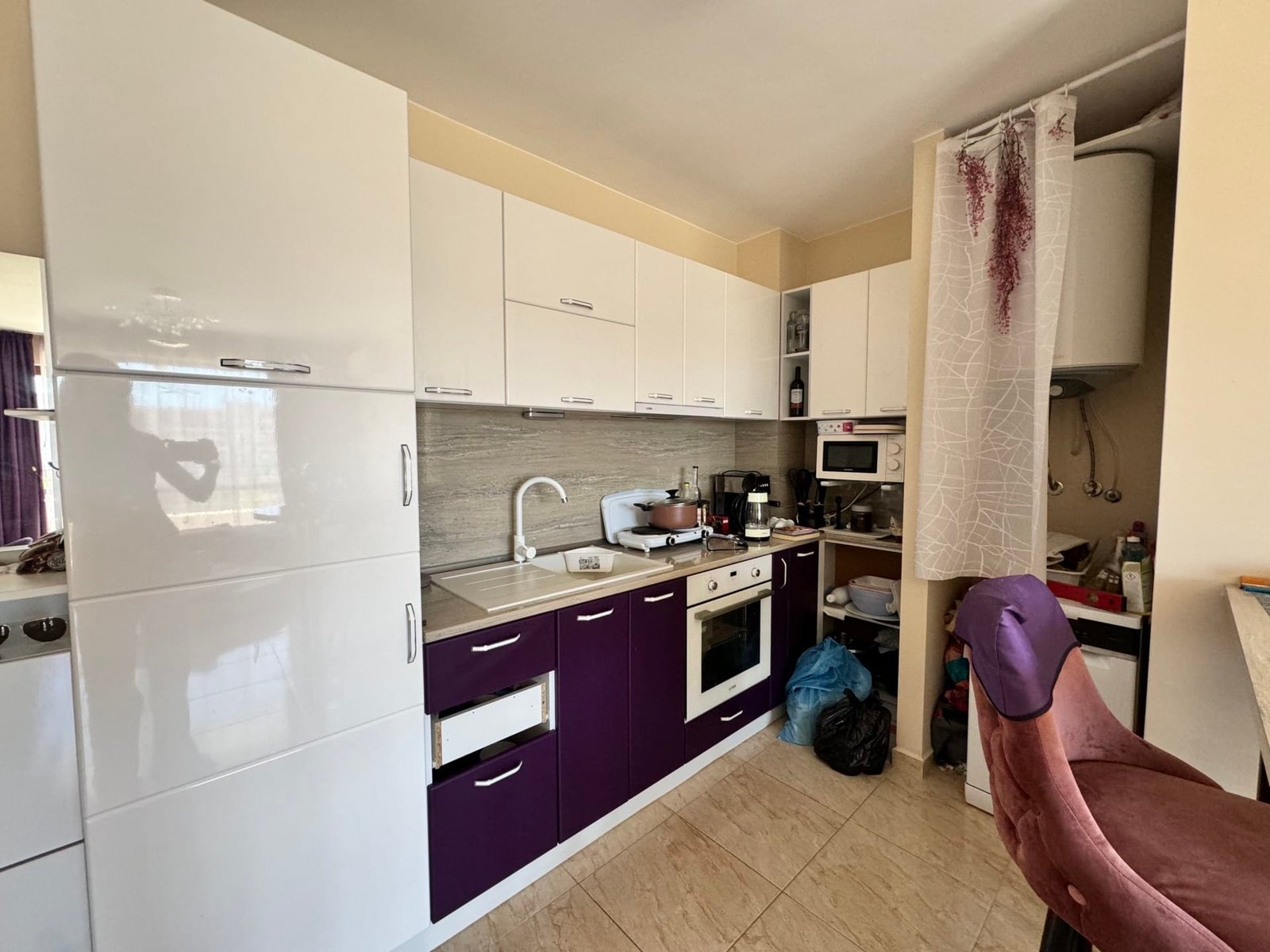 Современная кухня с бело-фиолетовой мебелью и техникой Современная кухня с белыми и фиолетовыми фасадами, компактная планировка, бытовая техника и обеденные стулья.