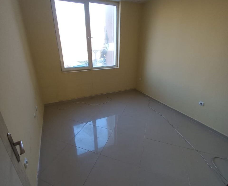 Светлая комната с желтыми стенами и большой окном, готовая к продаже или аренде в Болгарии. Полы выполнены из плитки.