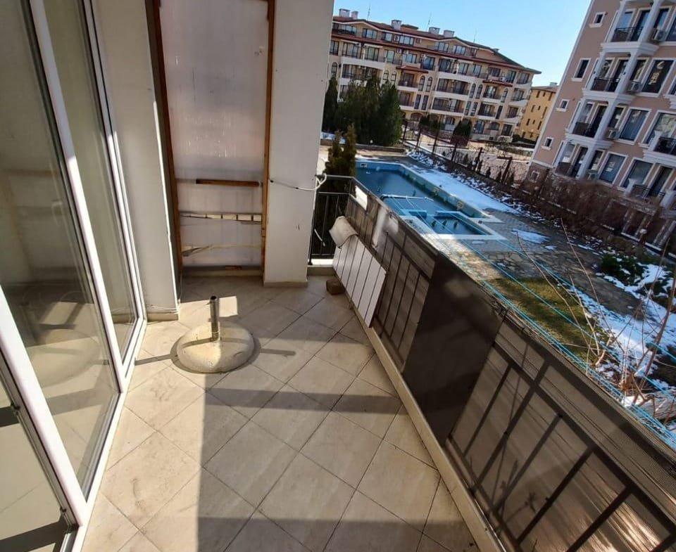 Балкон с плиточным полом и панорамным видом на бассейн в жилом комплексе в Болгарии. Чистое небо и зелень в окрестностях.