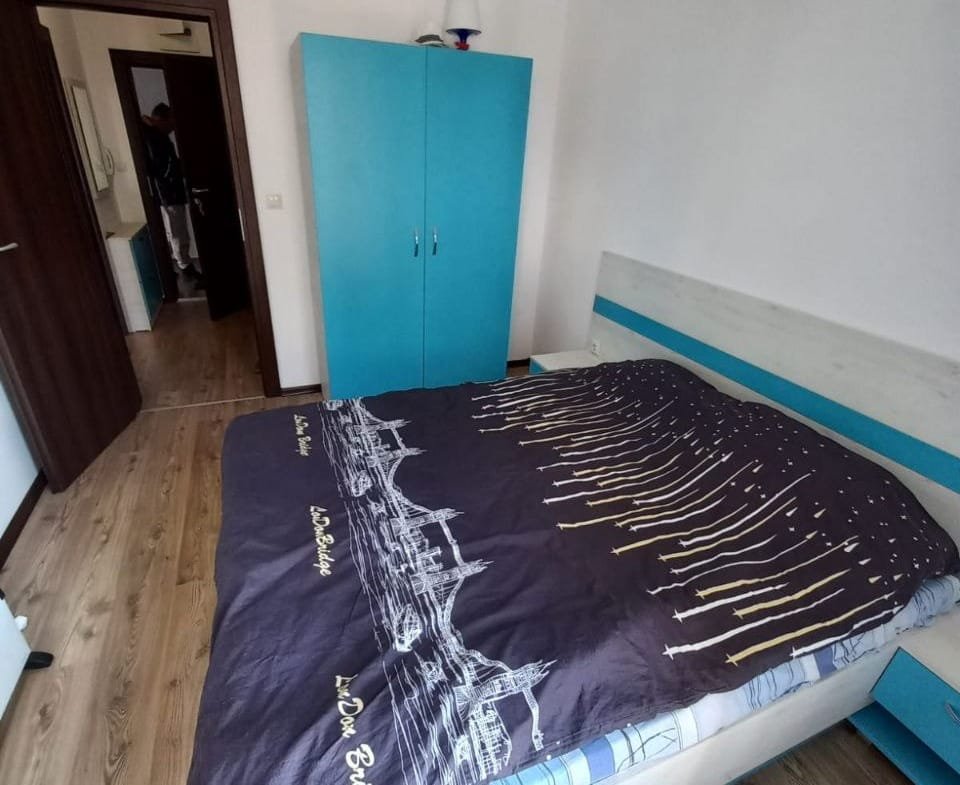 Современная спальня с двуспальной кроватью, голубым шкафом и светлым деревянным полом, предлагающая уют для проживания в Болгарии.