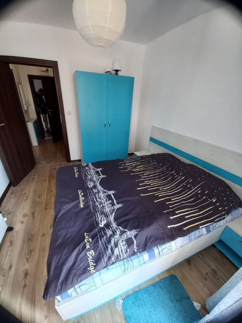 Уютная спальня с синим шкафом и удобной кроватью Современная спальня с двуспальной кроватью, голубым шкафом и светлым деревянным полом, предлагающая уют для проживания в Болгарии.