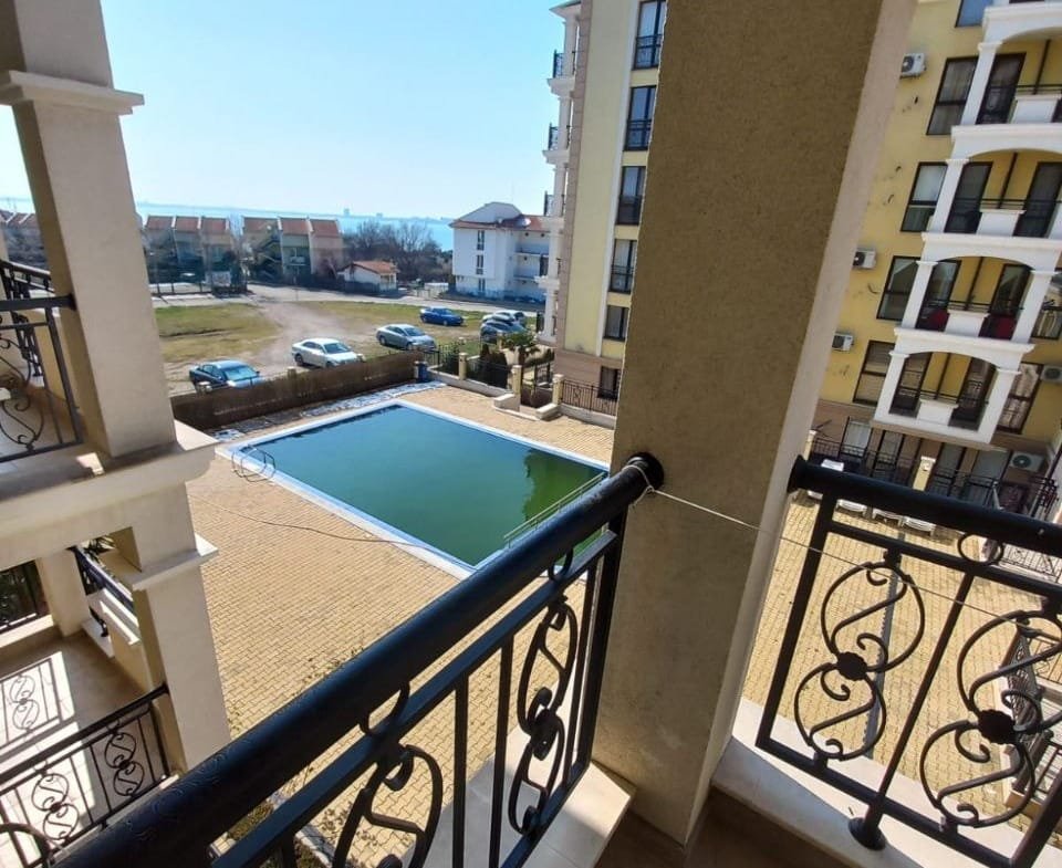 Балкон с видом на зелёный участок и бассейн в комплексе недвижимости в Болгарии, солнце светит ясно на пейзаже.