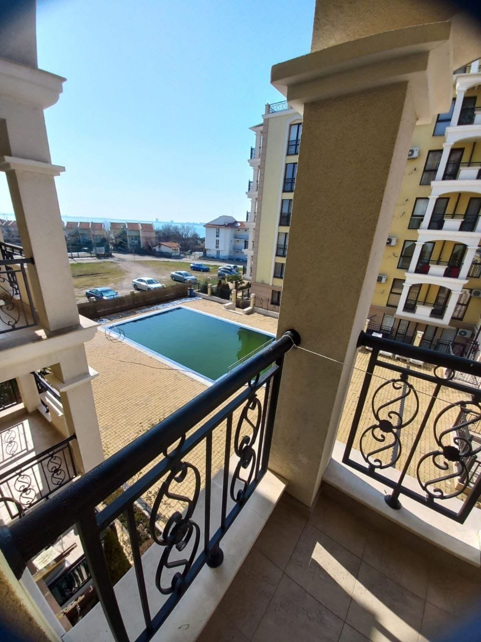 Балкон с видом на бассейн и жилой комплекс под солнечным небом Балкон с видом на зелёный участок и бассейн в комплексе недвижимости в Болгарии, солнце светит ясно на пейзаже.