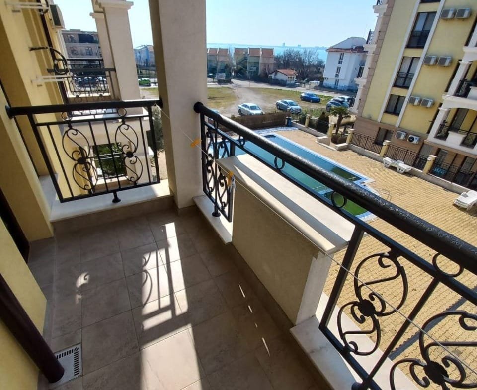 Балкон с решеткой, вид на бассейн и жилые здания, яркое солнечное освещение, привлекающее внимание к недвижимости в Болгарии.