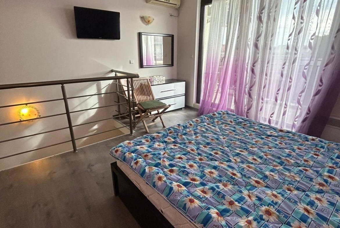 Светлая спальня с кроватью, телевизором, комодом и балконной дверью, оформленная в современном стиле для продажи в Болгарии.