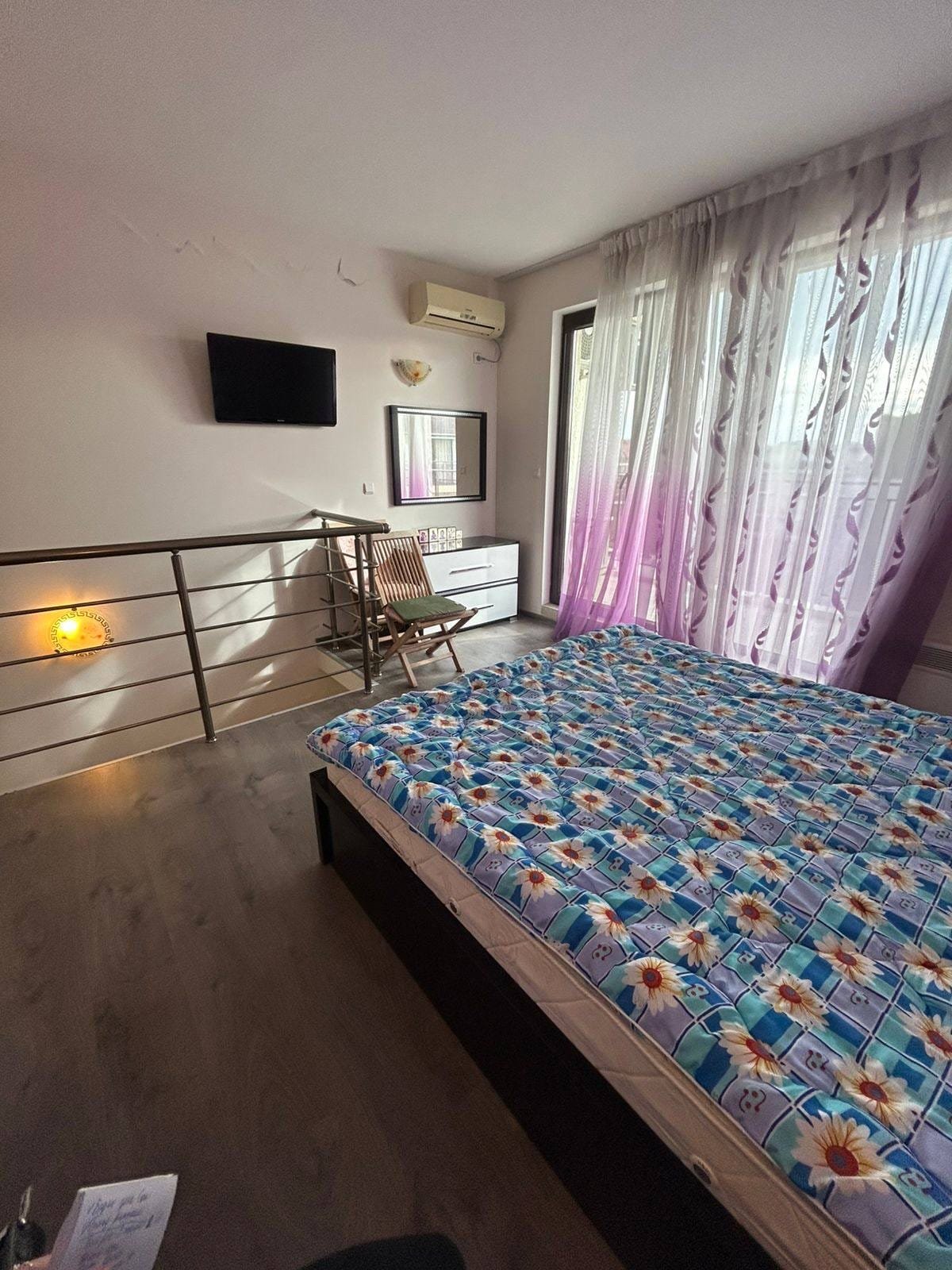 Современная спальня с большим окном и ярким текстилем Светлая спальня с кроватью, телевизором, комодом и балконной дверью, оформленная в современном стиле для продажи в Болгарии.