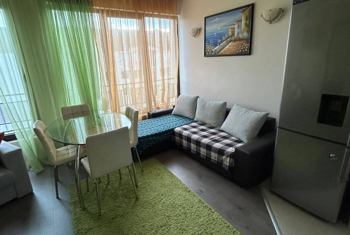 Современная квартира в Болгарии с уютной гостиной, большим окном, обеденным столом и холодильником. Зеленый ковер добавляет уют.