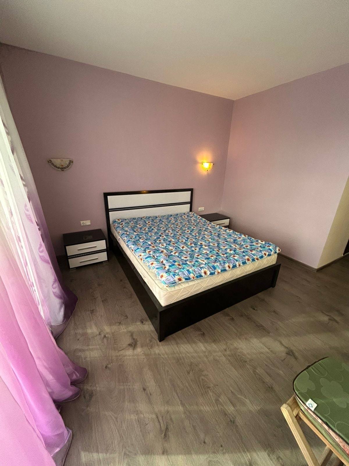 Современная спальня с кроватью и фиолетовыми стенами Современная спальня с кроватью, комодом и мягкими шторами пурпурного цвета, идеальна для аренды недвижимости в Болгарии.