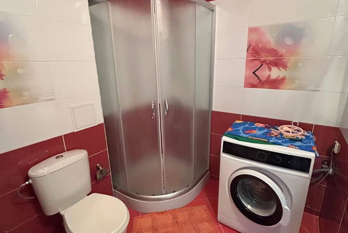 Современная ванная комната в Болгарии с душевой кабиной, унитазом и стиральной машиной на фоне красной плитки.