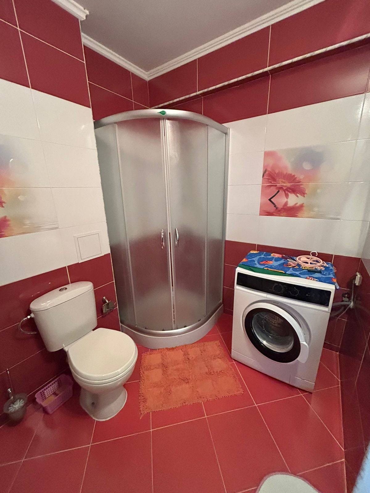 Современная ванная комната с душевой и стиральной машиной Современная ванная комната в Болгарии с душевой кабиной, унитазом и стиральной машиной на фоне красной плитки.