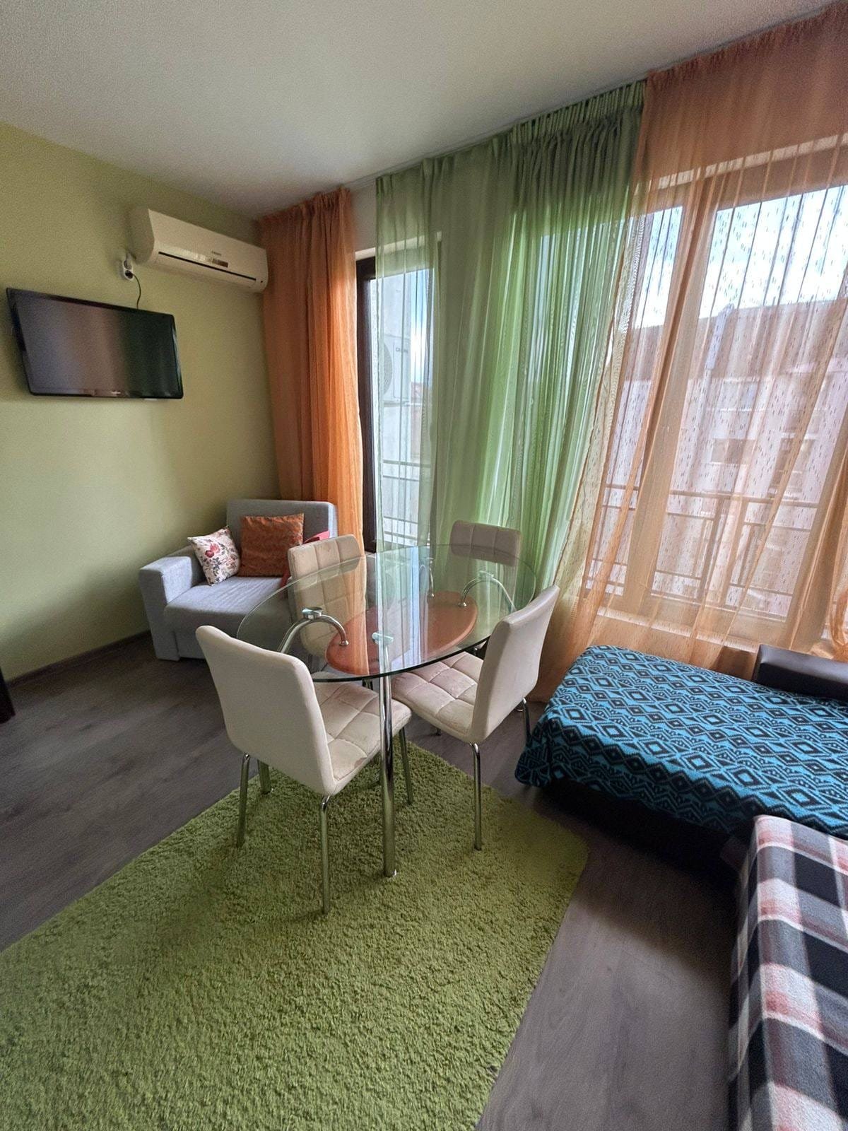 Современная квартира с обеденной зоной и светлыми шторами Современный интерьер квартиры в Болгарии: стол с прозрачными стульями, уютные диваны, яркие шторы и телевизор.