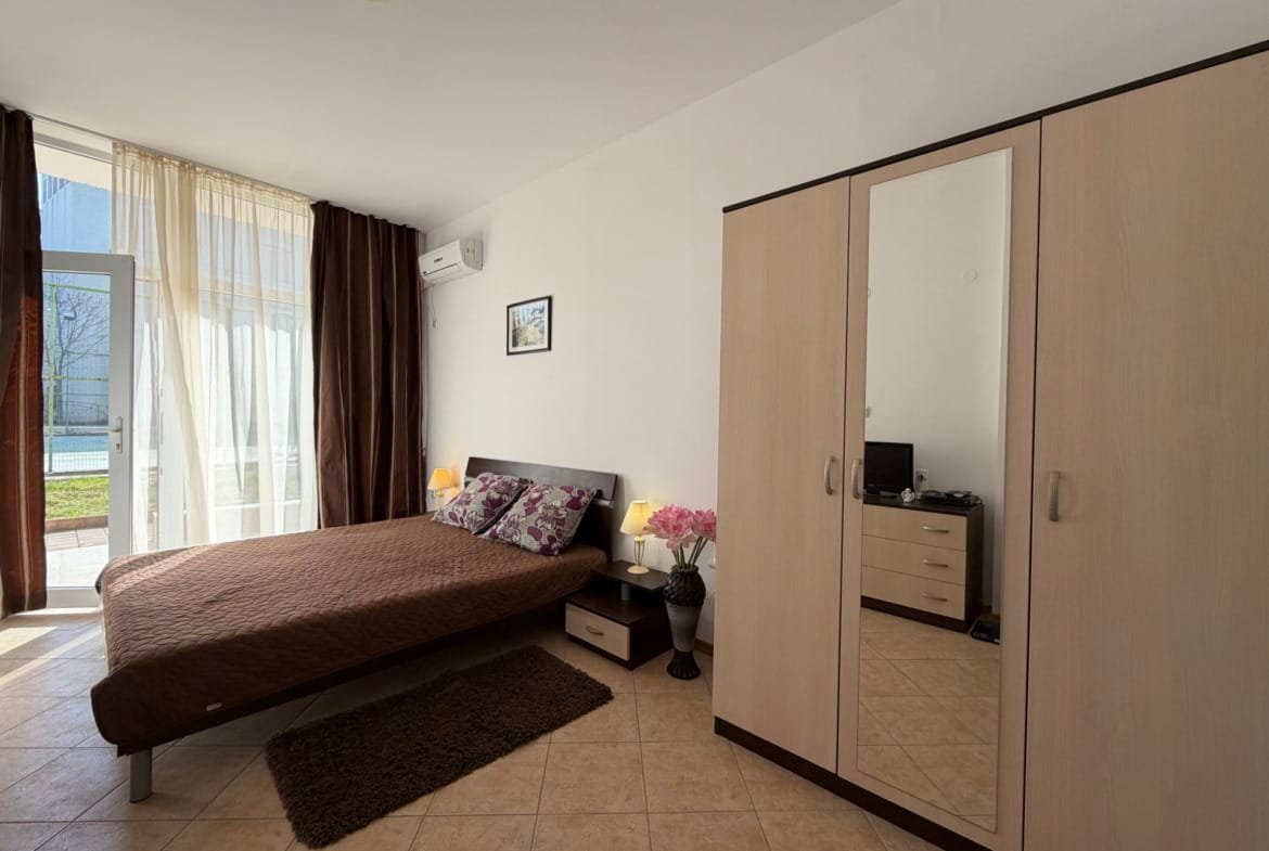 Уютная спальня с двуспальной кроватью, шкафом, мягким ковриком и светлыми шторами, подходящая для комфортного проживания в Болгарии.