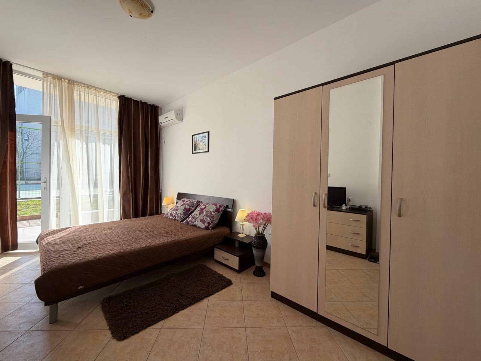 Уютная спальня с двуспальной кроватью, шкафом, мягким ковриком и светлыми шторами, подходящая для комфортного проживания в Болгарии.