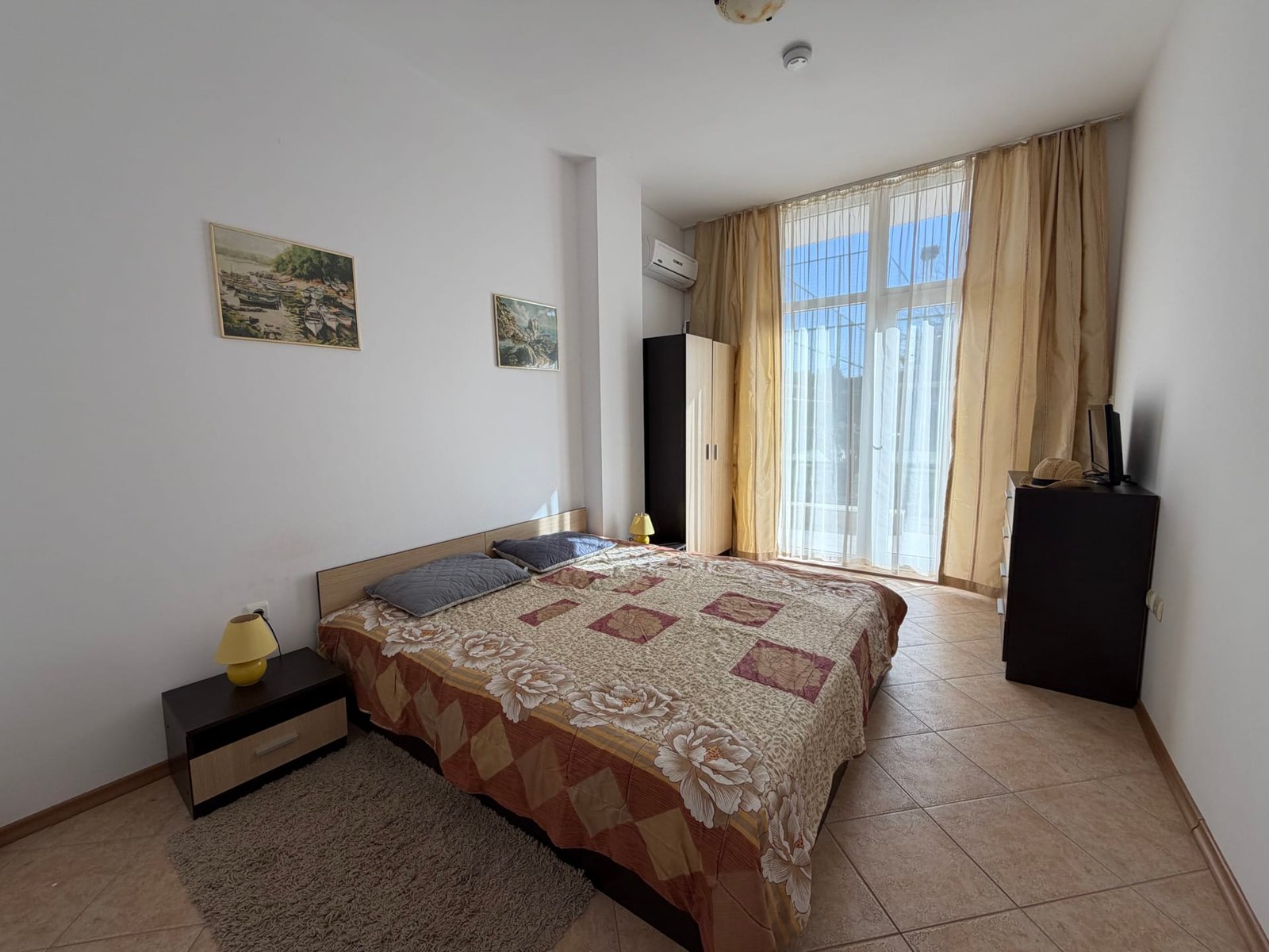 Современная спальня с двуспальной кроватью, светлыми шторами и выходом на балкон в квартире в Болгарии.