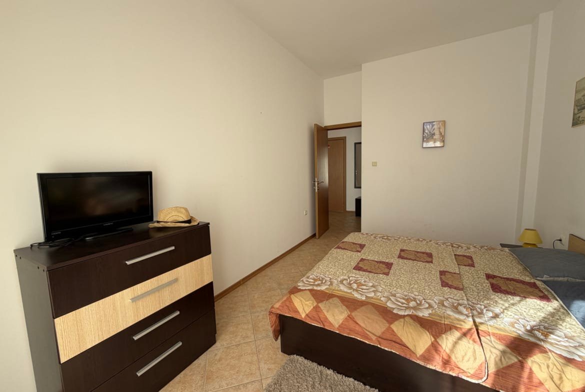 Современная спальня с кроватью, комодом, телевизором и нейтральными тонами, подходящая для комфортного проживания в Болгарии.