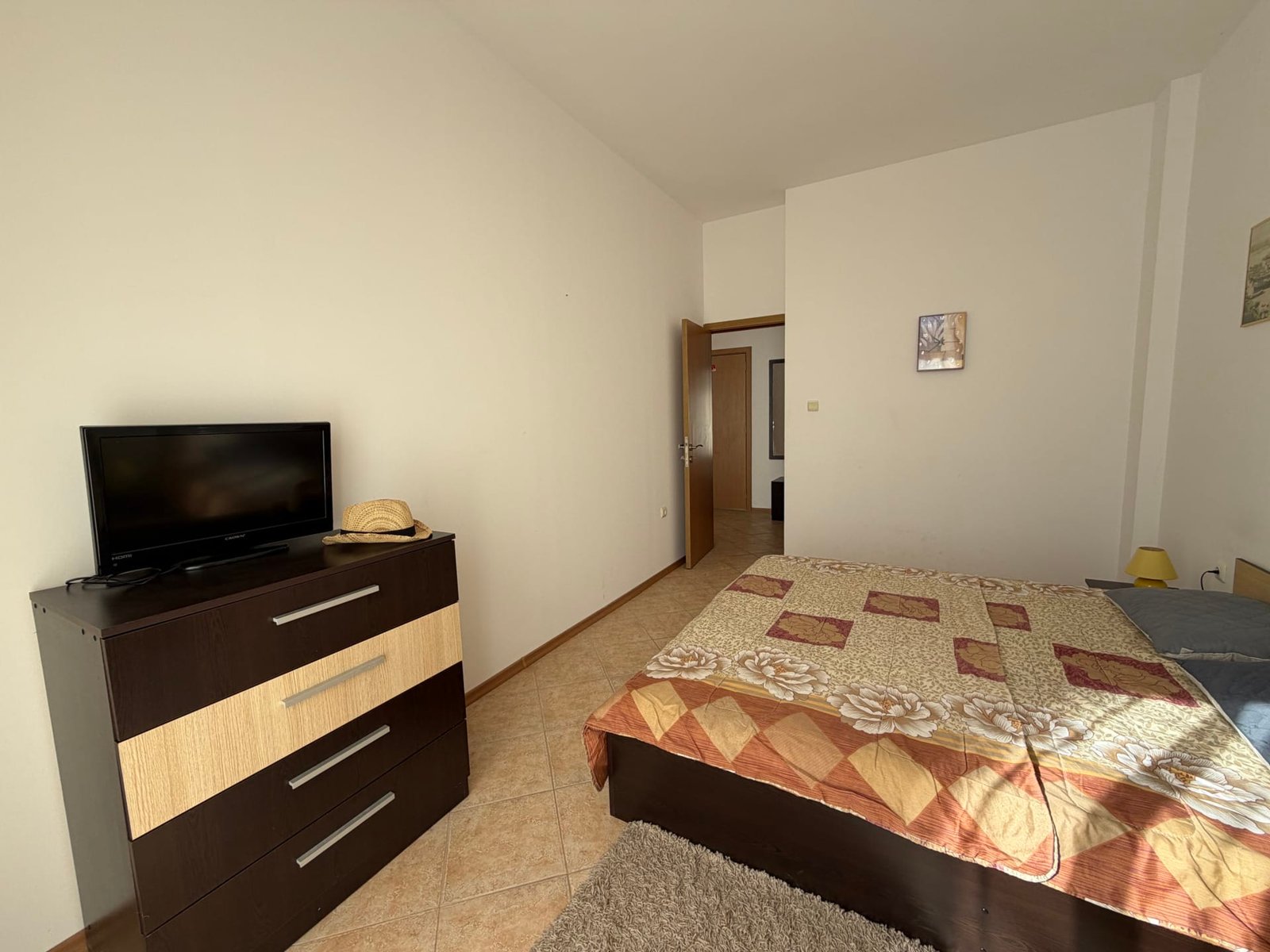 Современная спальня с кроватью, комодом, телевизором и нейтральными тонами, подходящая для комфортного проживания в Болгарии.