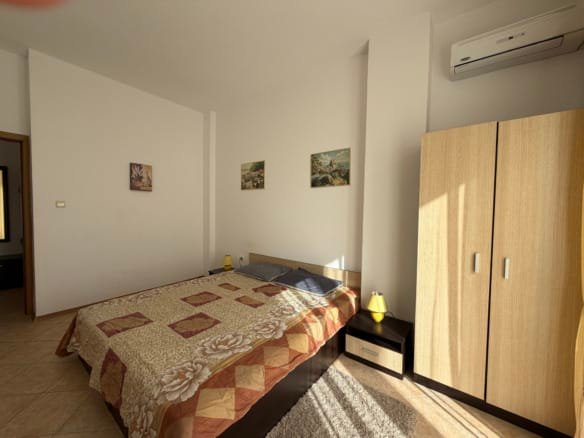 Светлая спальня с кроватью, деревянным шкафом и картинами на стенах, идеальна для отдыха в недвижимости Болгарии.