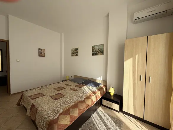 Светлая спальня с кроватью, деревянным шкафом и картинами на стенах, идеальна для отдыха в недвижимости Болгарии.