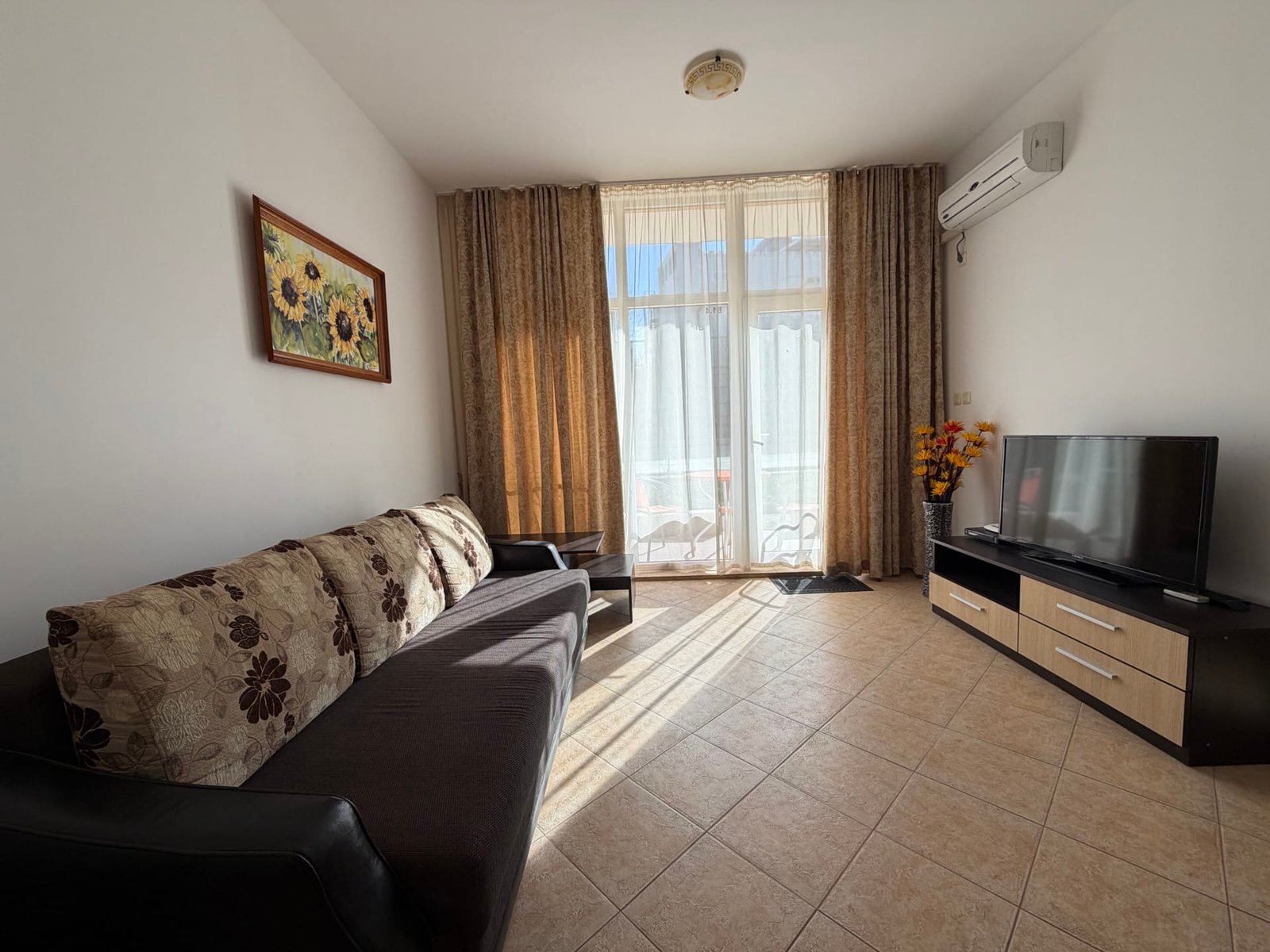 Солнечная гостиная с диваном и телевизором, окнами, выходящими на балкон, идеальна для покупки недвижимости в Болгарии.