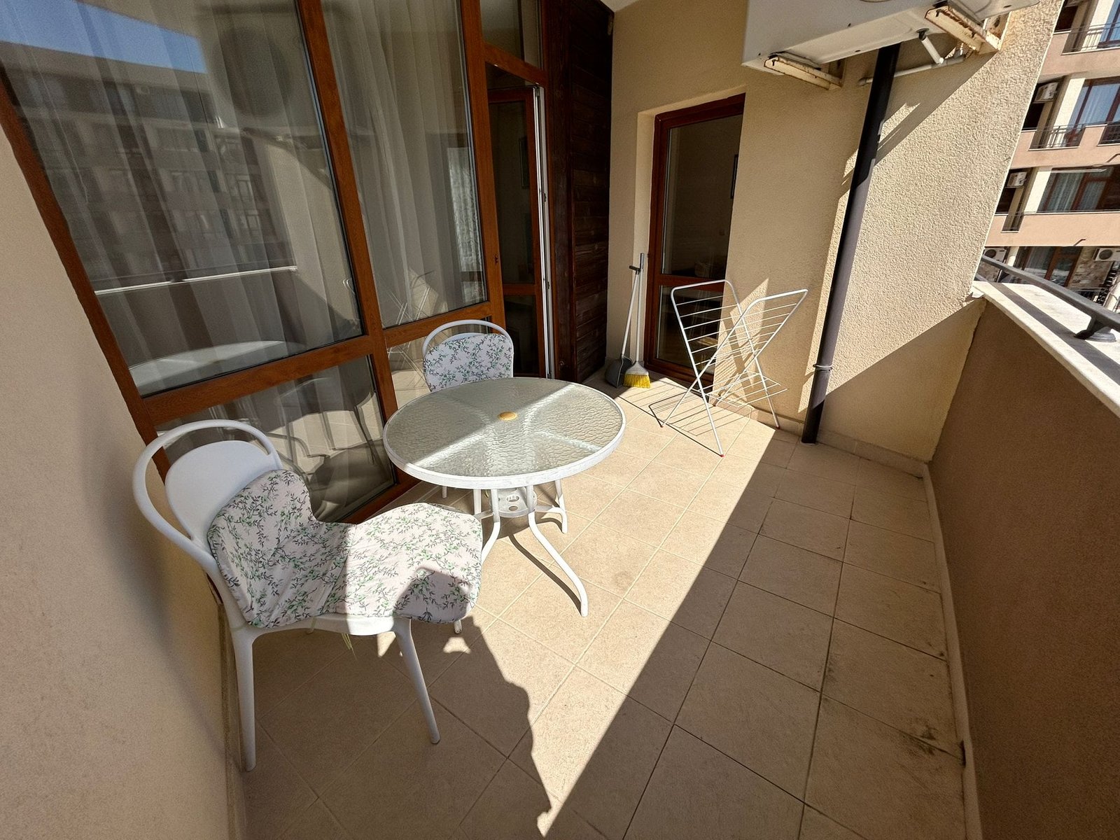 Уютный балкон с обеденным столом и стульями на солнечном свете. Балкон с круглым столом и стульями, на полу плитка, освещён солнечным светом, идеальное место для отдыха в Болгарии.