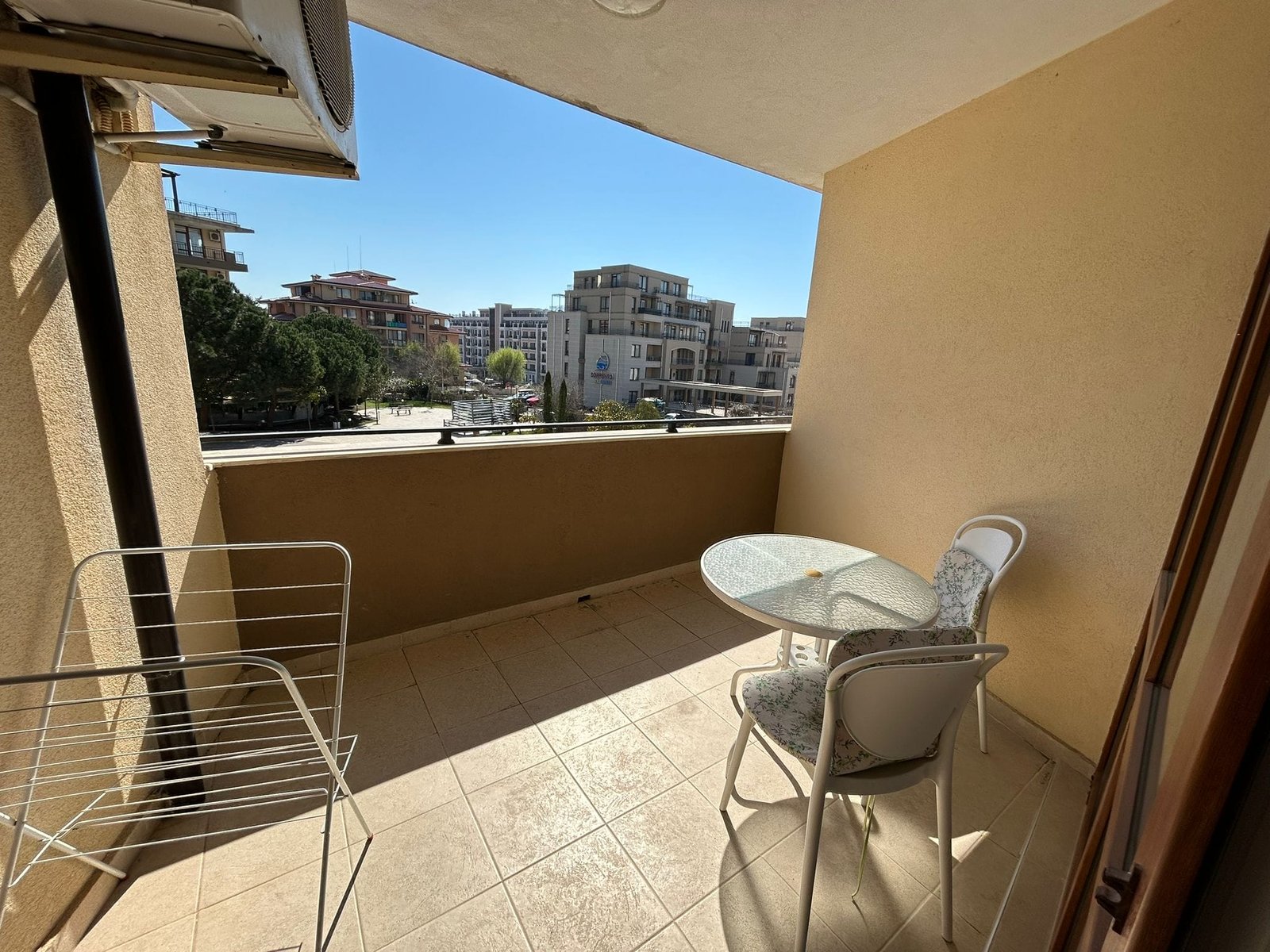 Балкон с обеденной зоной и видом на город в солнечный день Балкон с круглым стеклянным столом и стульями, вид на городские здания в Болгарии, идеальное место для отдыха.