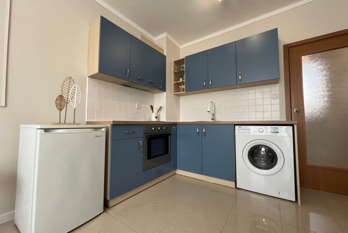 Современная кухня в квартире с синими фасадами, оборудована плитой, раковиной и стиральной машиной, идеальна для жизни в Болгарии.