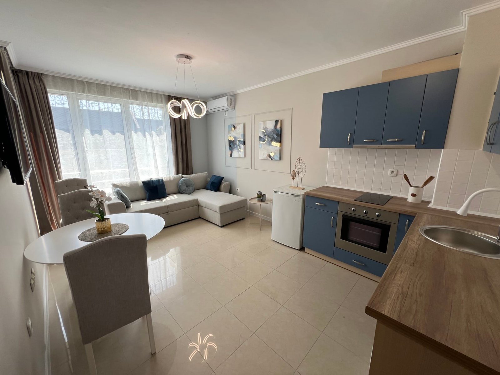 Современный интерьер квартиры в Болгарии с уютной гостиной, кухонным блоком, светлыми тонами и большими окнами.