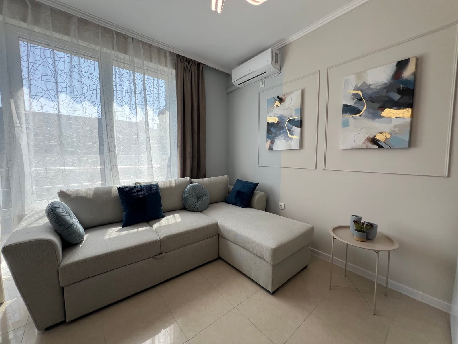 Современный уютный интерьер квартиры в Болгарии с диваном, светлыми стенами и декоративными картинами.