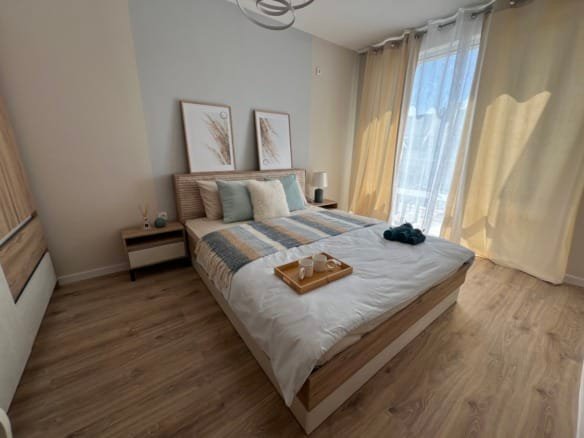 Современная спальня с уютной кроватью, светлыми шторами и деревянным полом, идеальна для покупки недвижимости в Болгарии.