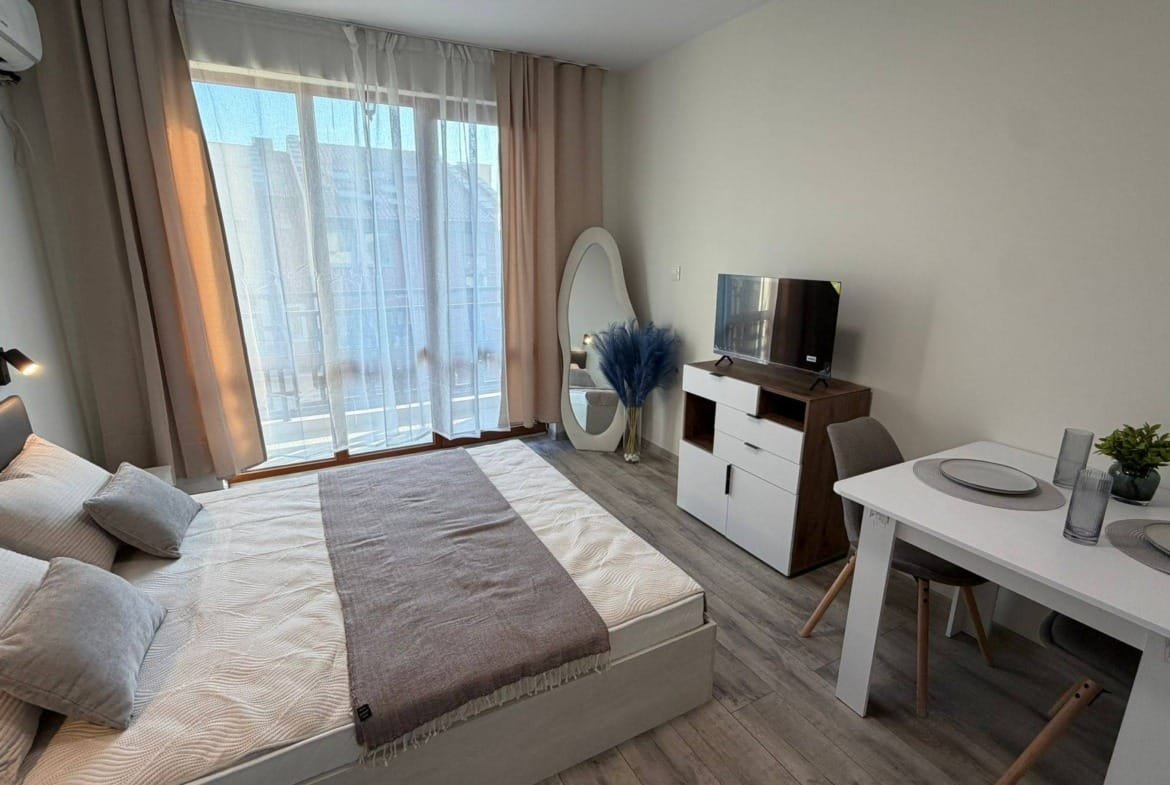 Светлая комната с двуспальной кроватью, телевизором и столом, идеально подходящая для покупки или аренды недвижимости в Болгарии.