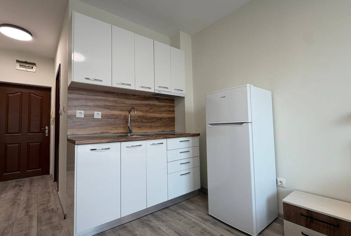 Современная кухня с белыми шкафами и деревянной столешницей, холодильником и моечными принадлежностями в квартире в Болгарии.