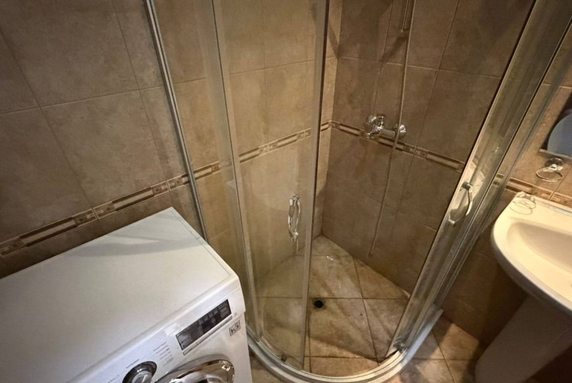 Душевая кабина с прозрачными стенками, рядом стиральная машина и умывальник в ванной комнате недвижимости в Болгарии.