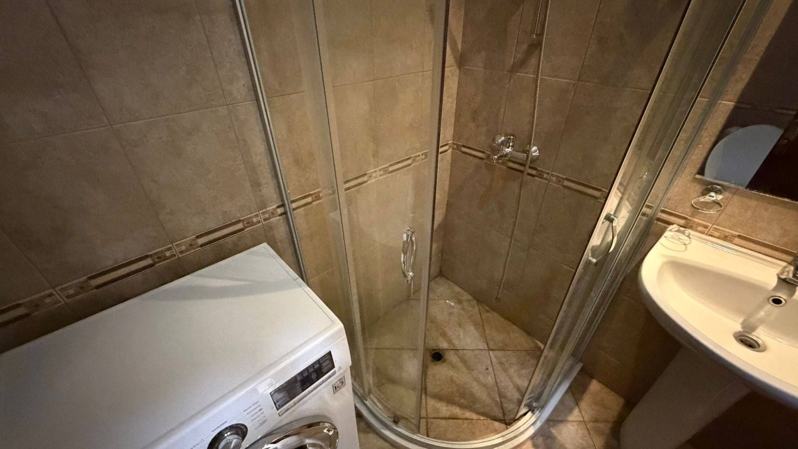 Душевая кабина с прозрачными стенками, рядом стиральная машина и умывальник в ванной комнате недвижимости в Болгарии.