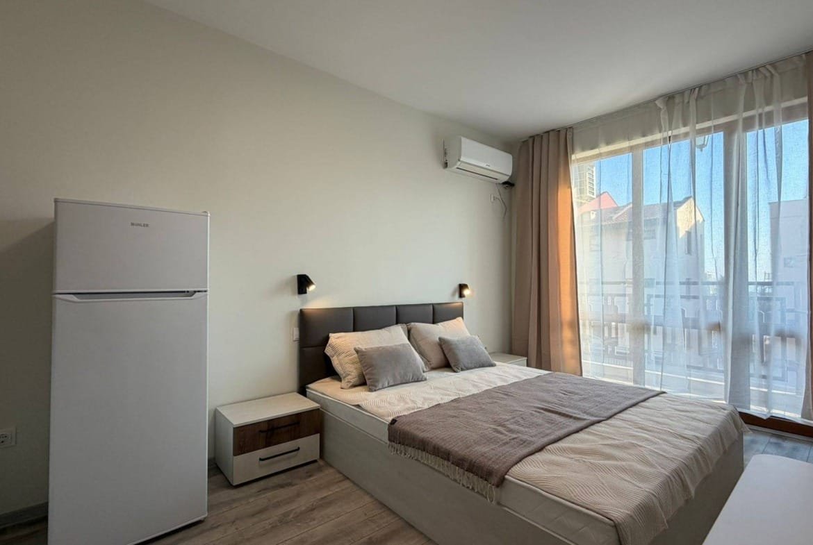 Современная спальня с белой мебелью, большим окном и балконом, готовая для аренды или покупки в Болгарии.