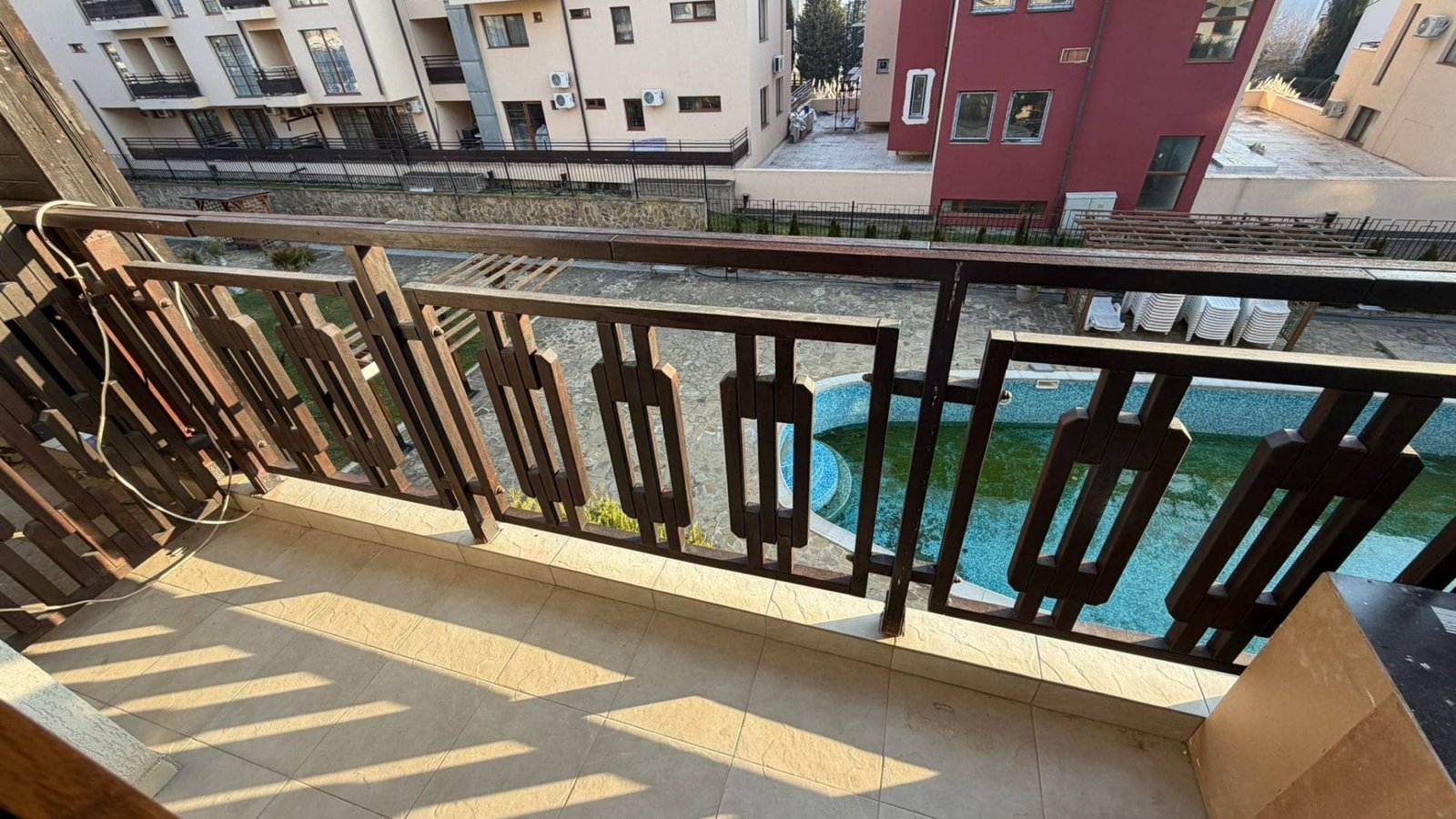 Балкон с деревянной оградой, солнечное освещение, вид на двор с бассейном и зданиями, подходящими для покупки недвижимости в Болгарии.