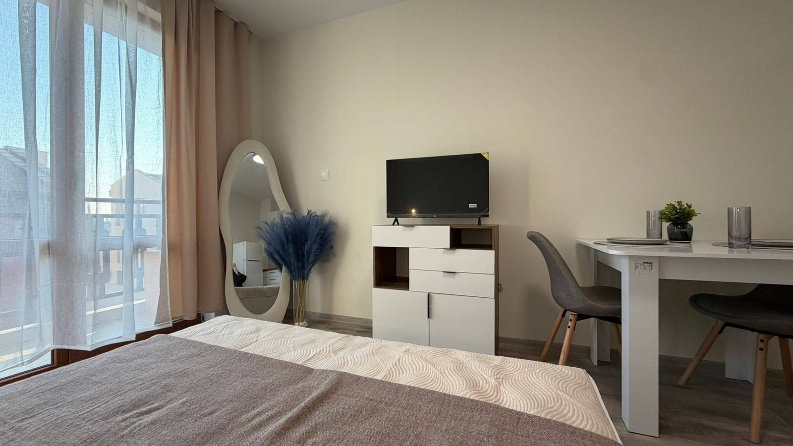 Современная квартира в Болгарии с теплым интерьером, телевизором, столом и зеркалом, идеальная для аренды или покупки.