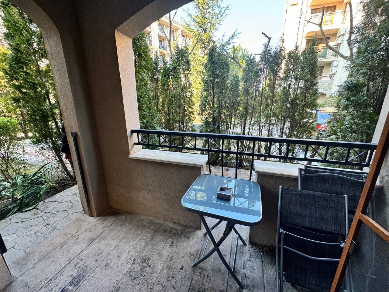 Балкон с небольшой столик и стульями, окружённый зелёными растениями, идеальное место для отдыха при продаже недвижимости в Болгарии.