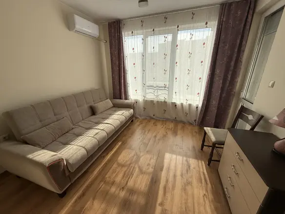 Комфортная гостиная с диваном, деревянным полом и большими окнами, оформлена в светлых тонах для продажи в Болгарии.